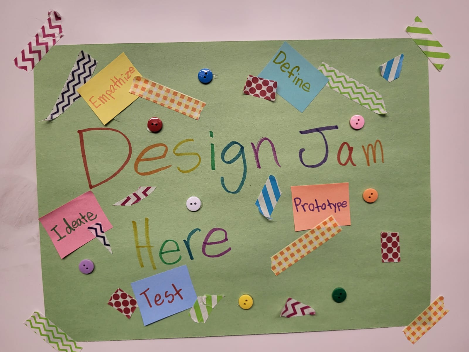 A hand-crafted sign that says Design Jam Here written in multi-colour on a green piece of paper.